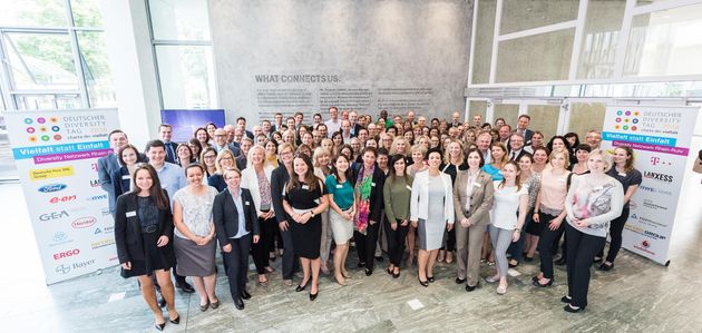 Gruppenfoto mit ca. 60 Personen des Diversity Netzwerk Rhein-Ruhr in einem hohen und hellen Raum. Rechts und links der Gruppe stehen Aufsteller mit den Logos der teilnehmenden Unternehmen.
