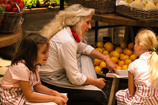 Eine Erwachsene erklärt zwei Mädchen etwas vor gefüllten Obstkörben