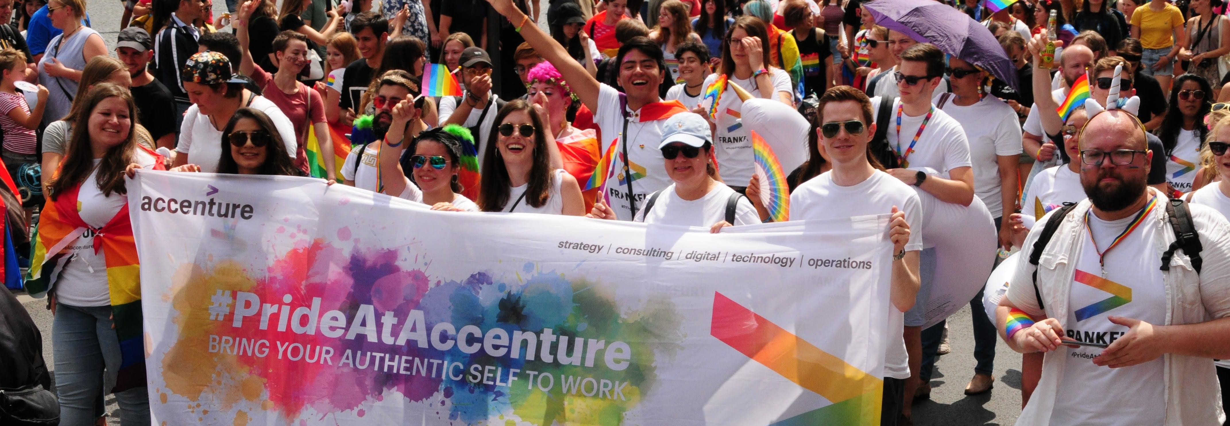 Auf dem Bild sind viele Personen zu sehen, die gemeinsam ein Banner halten auf dem #PrideAtAccenture steht. Sie tragen weiße Kleidung, passende T-Shirts und haben zum Teil Schilder oder Regenbogenfahnen in der Hand. 