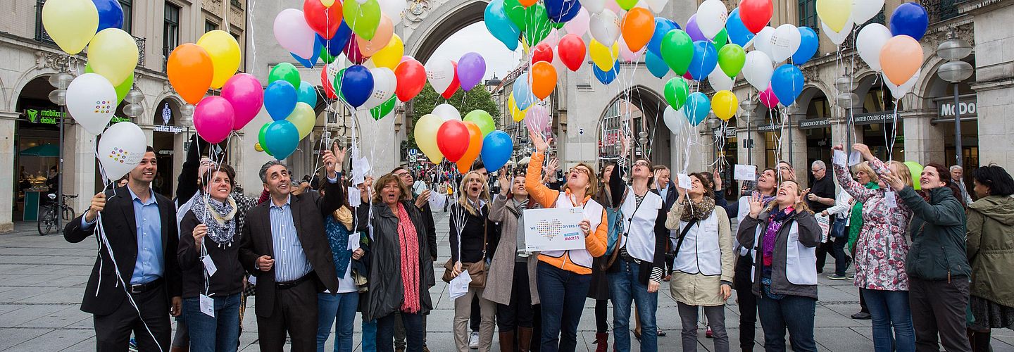 [Translate to Leichte Sprache:] Eine Gruppe Menschen lässt bunte Luftballons steigen