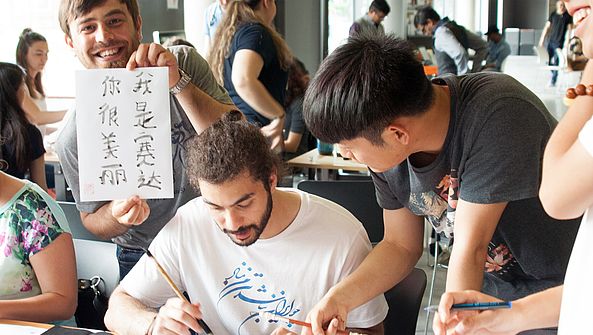 Mehrere Menschen stehen an einem Tisch und beugen sich zum Blatt vor, ein Mann hält ein Blatt mit chinesischer Kalligraphie und lächelt in die Kamera.