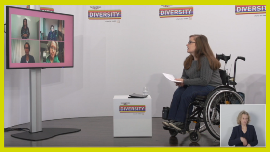 Zu sehen ist eine Moderatorin im Rollstuhl im Konferenz-Studio. Auf einem Bildschirm sind vier Gesprächsteilnehmende eingeblendet. Die Moderatorin ist dem Bildschirm zugewandt.